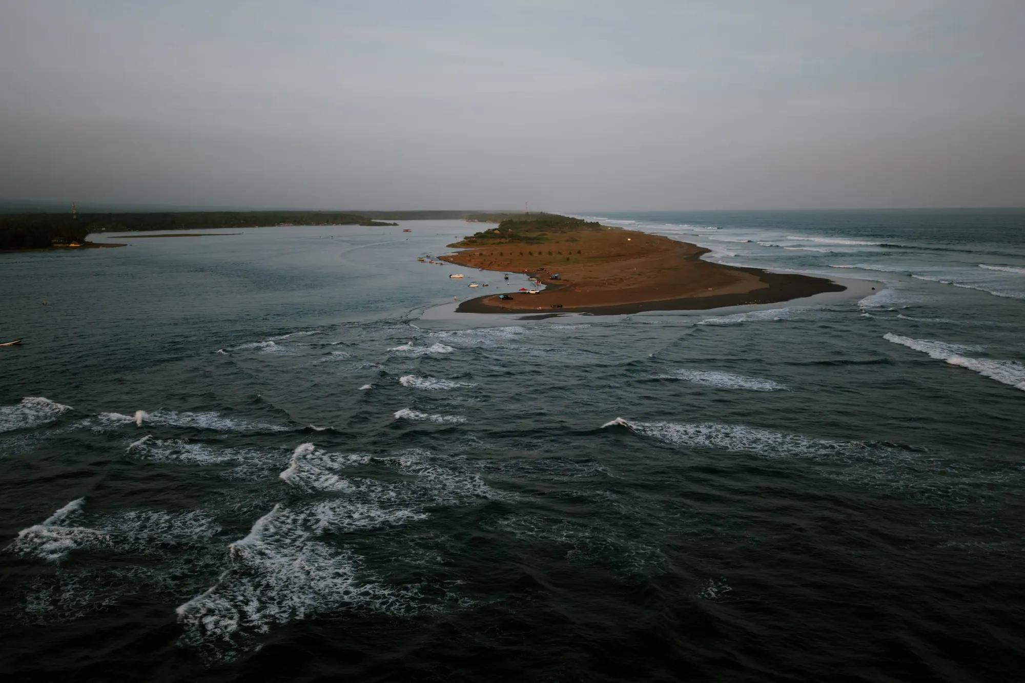 An aerial view of the sandbar tip at the river mouth, with pangas and vehicles on the sand, the flat estuary to one side and Pacific swell wrapping around the point on the other