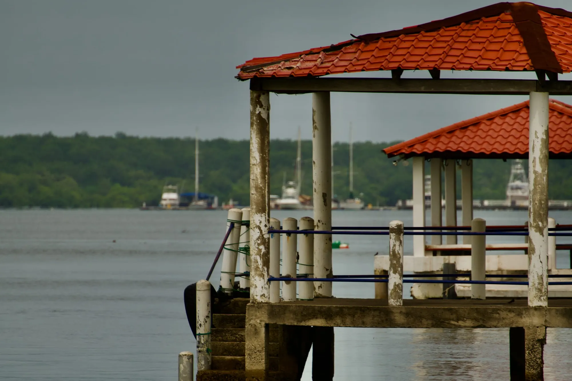 A concrete pier with orange tile-roofed shelters over flat estuary water, with moored boats and a mangrove treeline behind