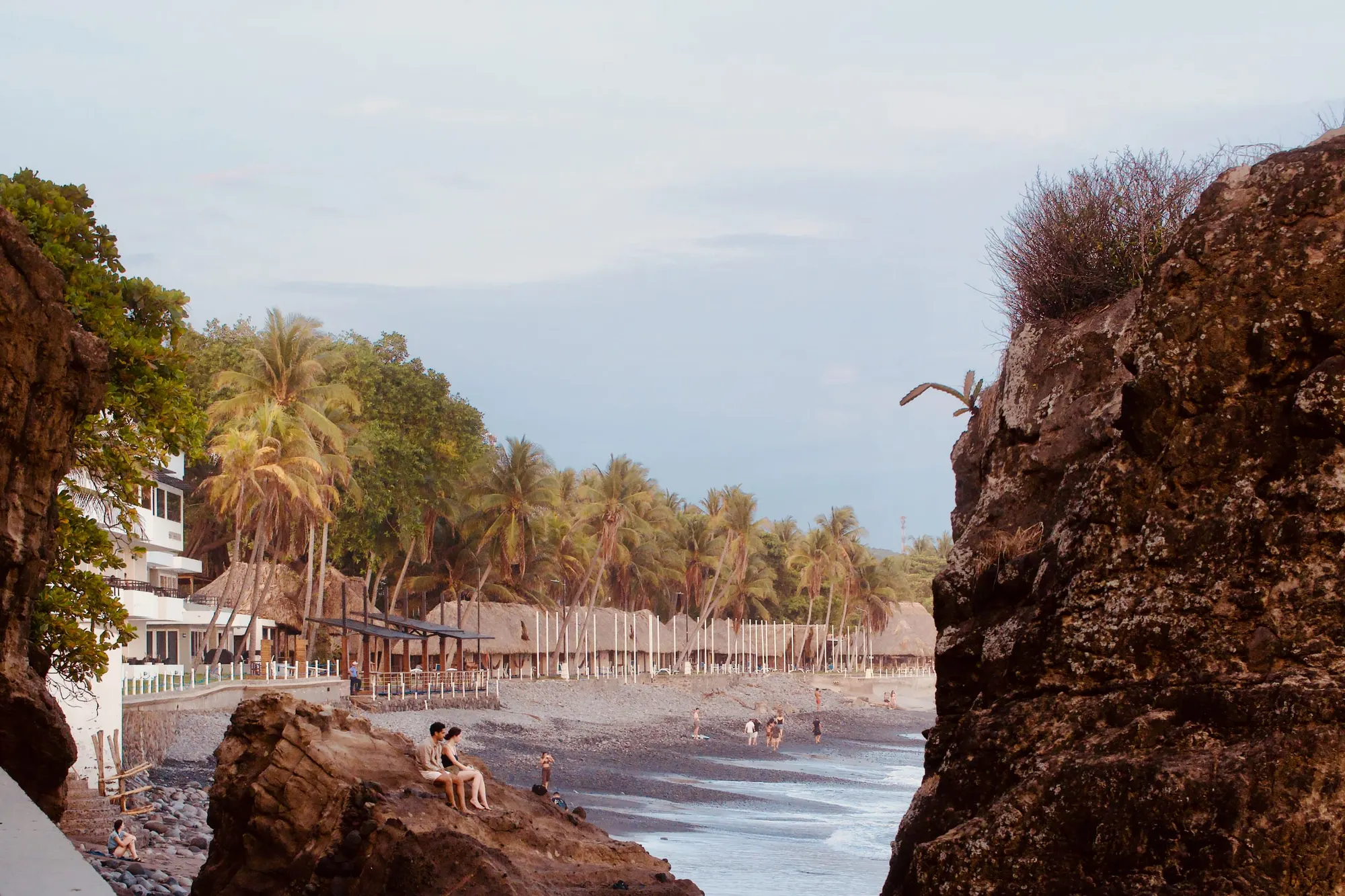 Palm trees and thatched beachfront restaurants along a dark cobble beach, framed between two large rock outcrops, with people sitting on a boulder in the foreground