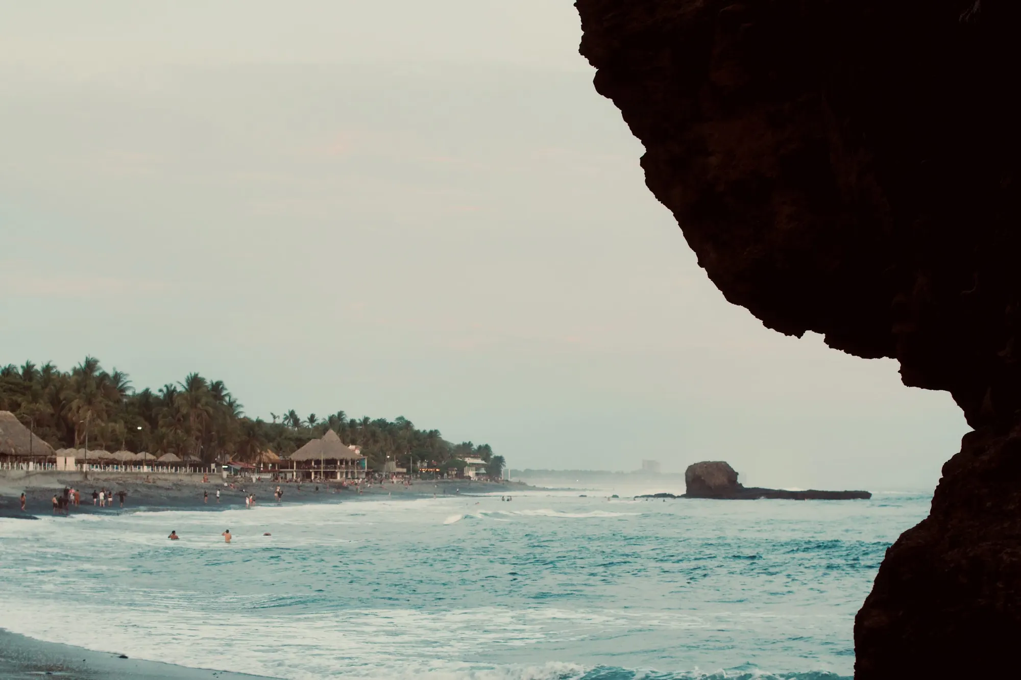 A dark sand beach curving away past palm trees and thatched roofs, with a detached rock stack standing offshore in the haze