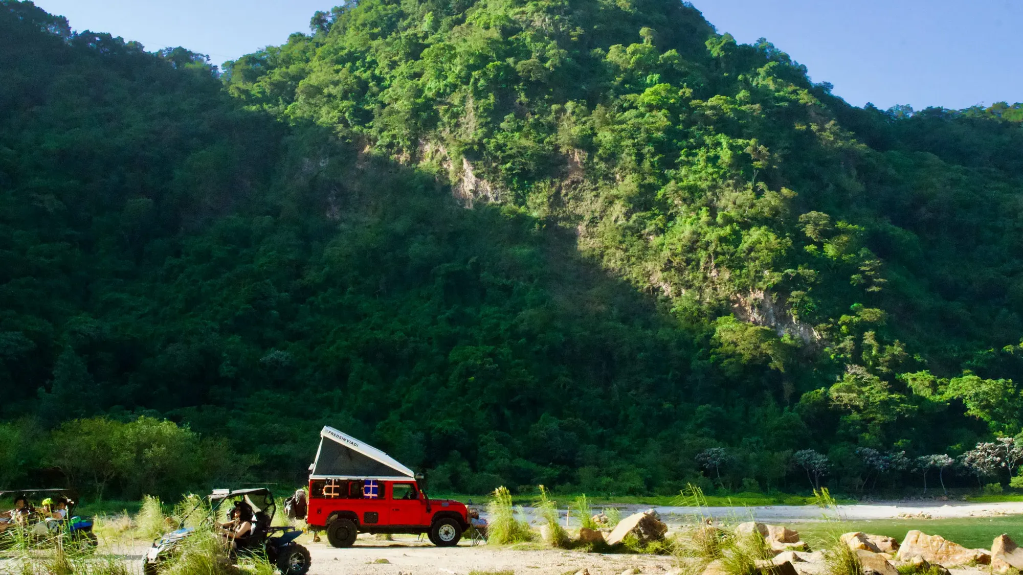 A red Land Rover Defender with its pop-top roof tent open, FREDSINVIADI printed along the tent, parked on a pale gravel flat below a steep forested hillside, with camp chairs, a side-by-side and a quad alongside it