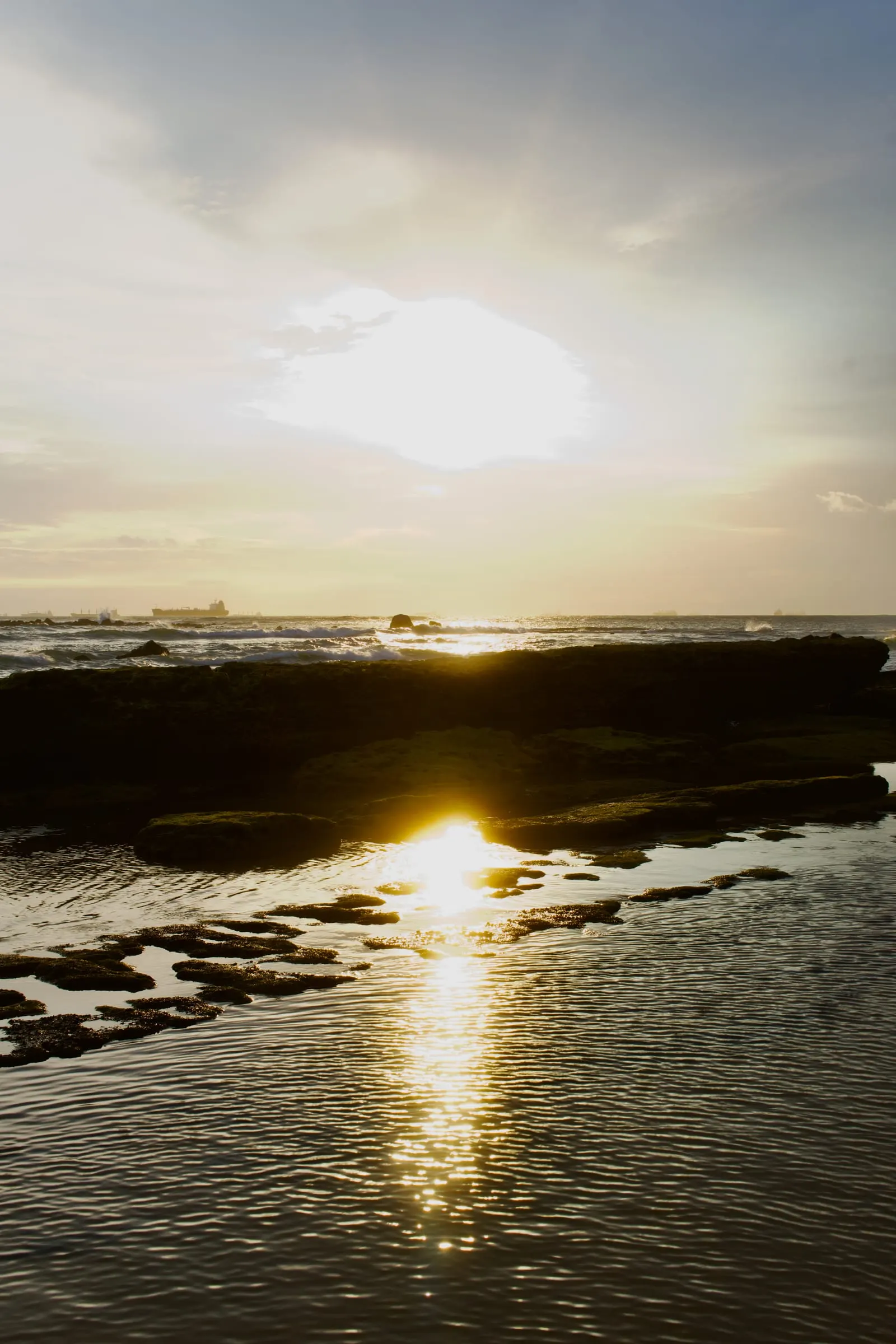 Low sun setting over a flat volcanic rock shelf and tide pools, with cargo ships anchored along the horizon